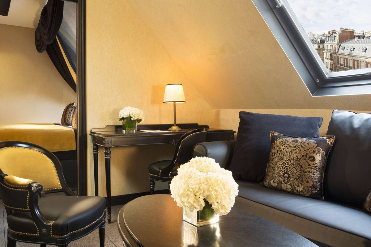 A modern living room with a sofa, a chair, a small table with a vase of flowers, a lamp, and a view of the city through a skylight.