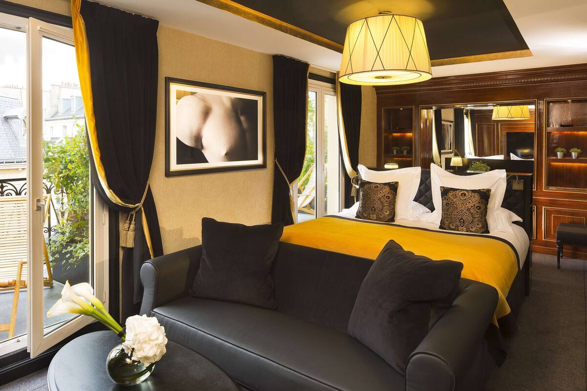 A hotel room with a large bed, a sofa, a dining area, and a view of the outside.