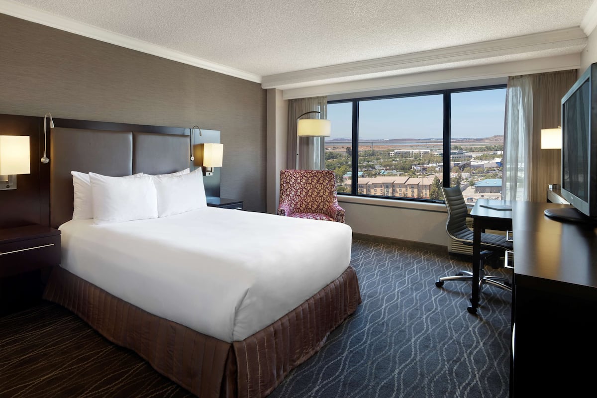 A hotel room with a large bed, a desk, a chair, and a view of the city through the window.
