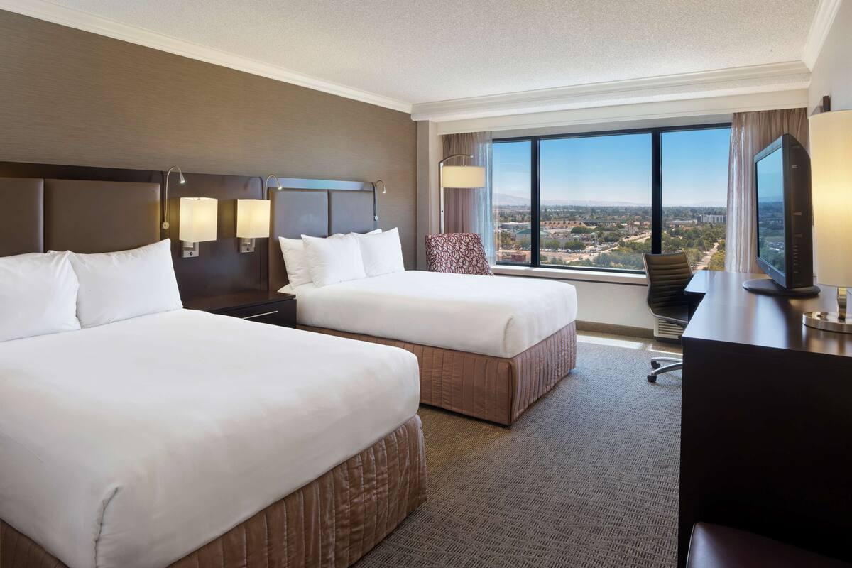 A hotel room with two beds, a desk, a chair, and a large window offering a city view.