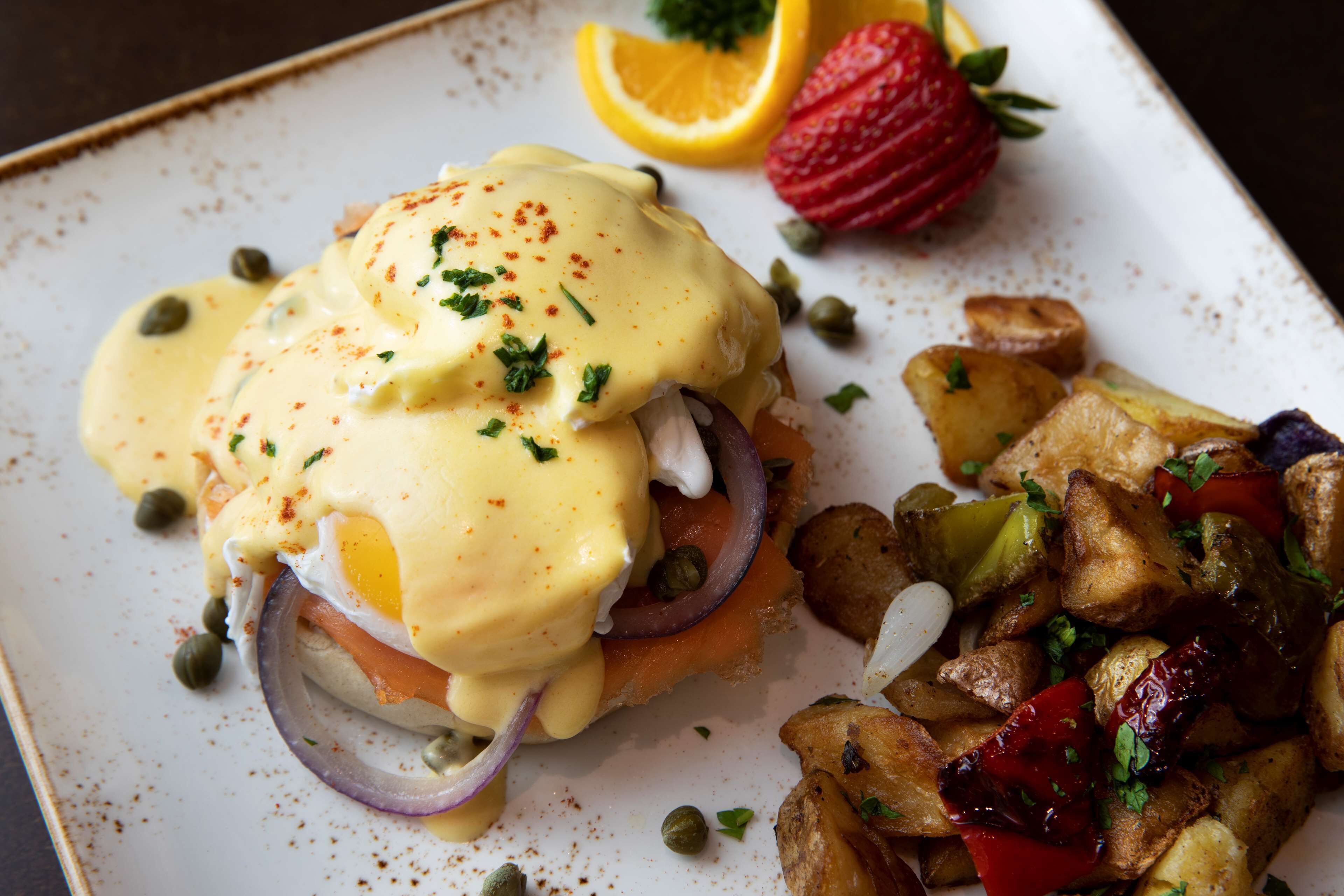 A plate with eggs benedict, capers, red onion, and a side of roasted potatoes and peppers.