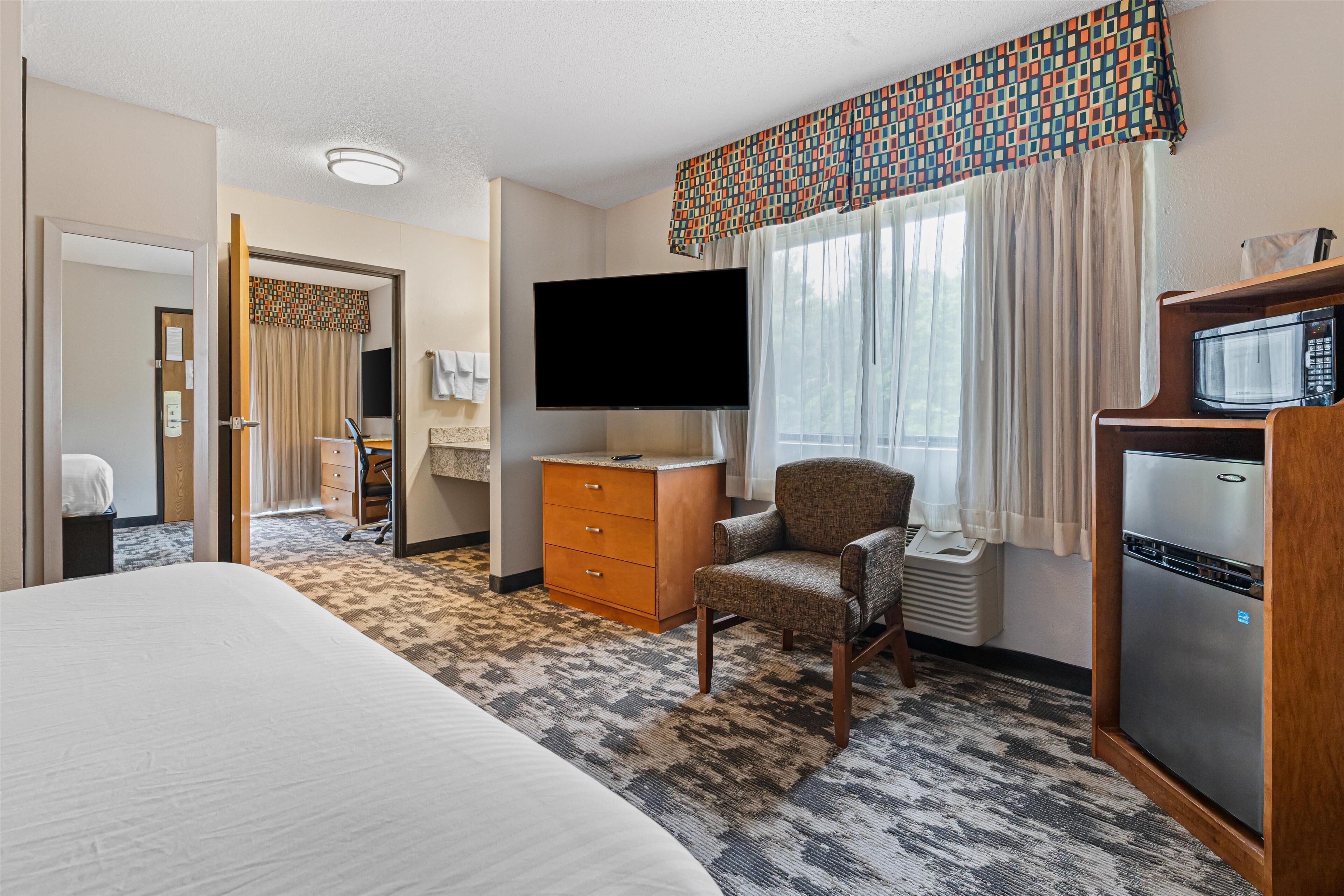 A hotel room with a large bed, a television, a refrigerator, a chair, a desk, and a window with curtains.