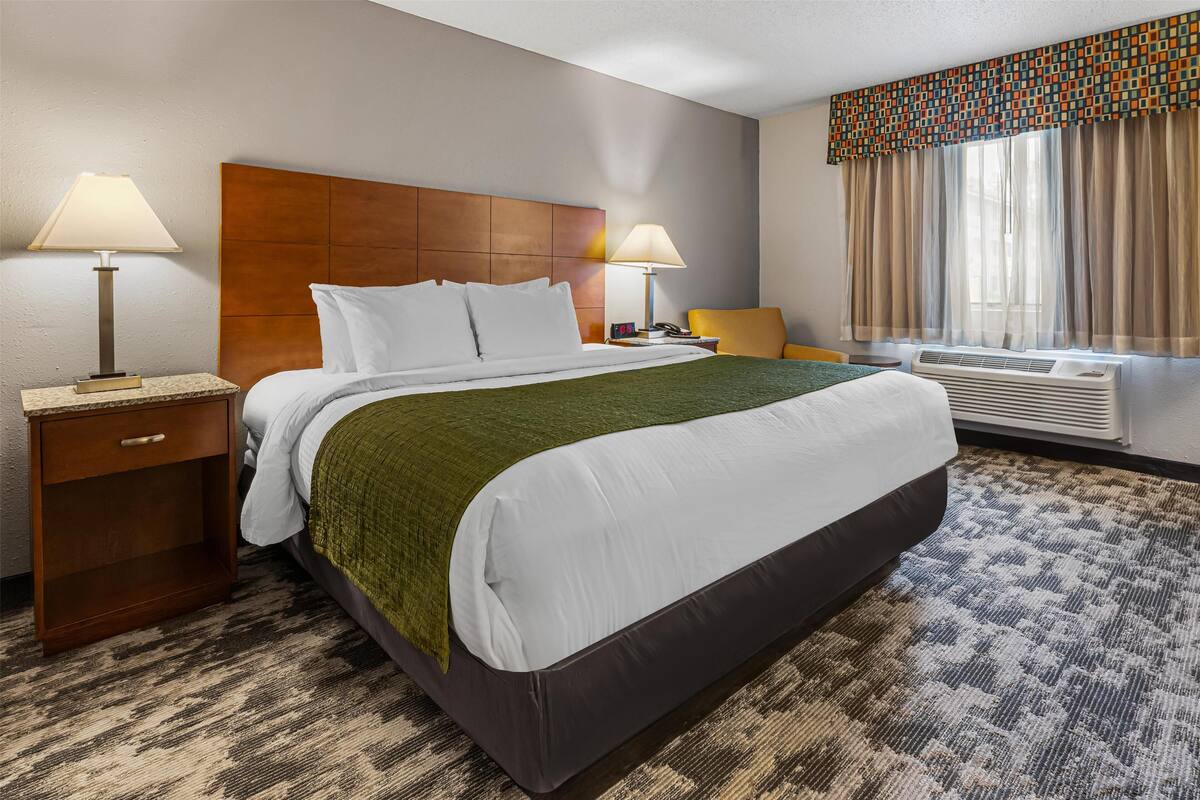A hotel room with a large bed, two bedside tables with lamps, a chair, a window with curtains, and a wall-mounted air conditioner.