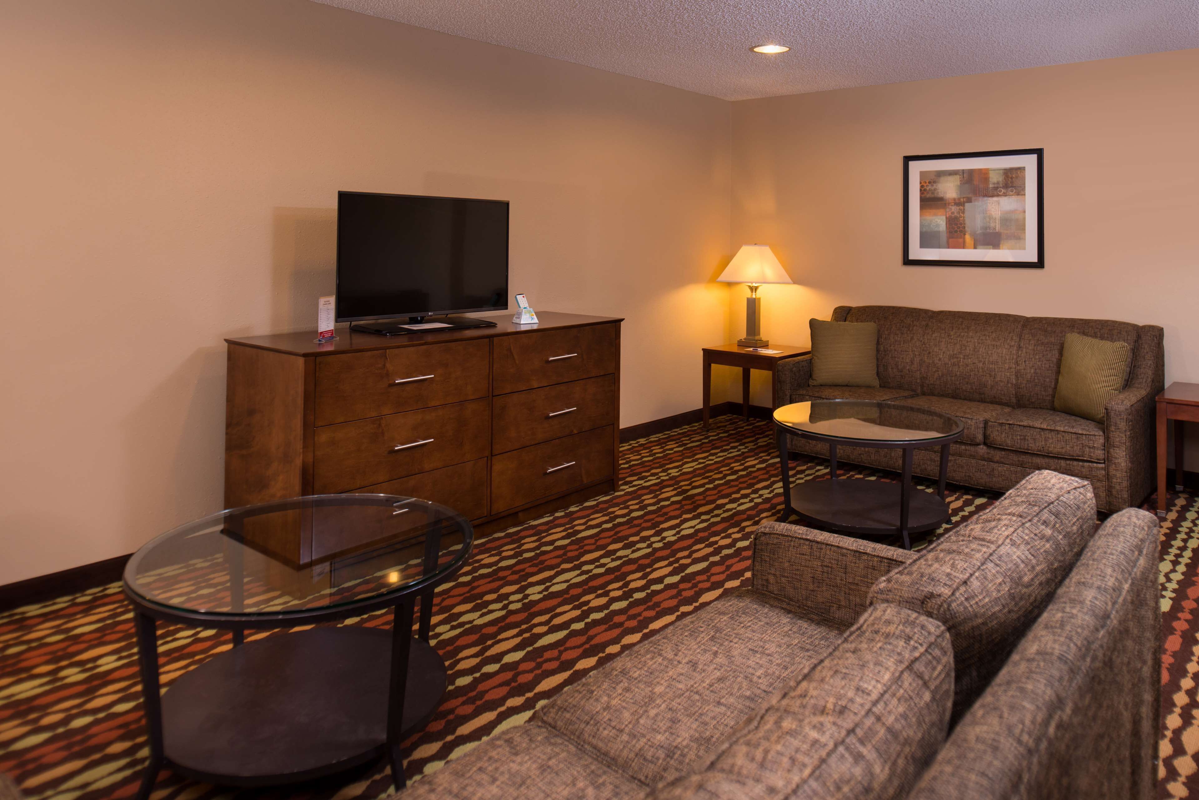 A hotel room with a sofa, a glass coffee table, a TV on a wooden cabinet, a lamp, and a patterned carpet.