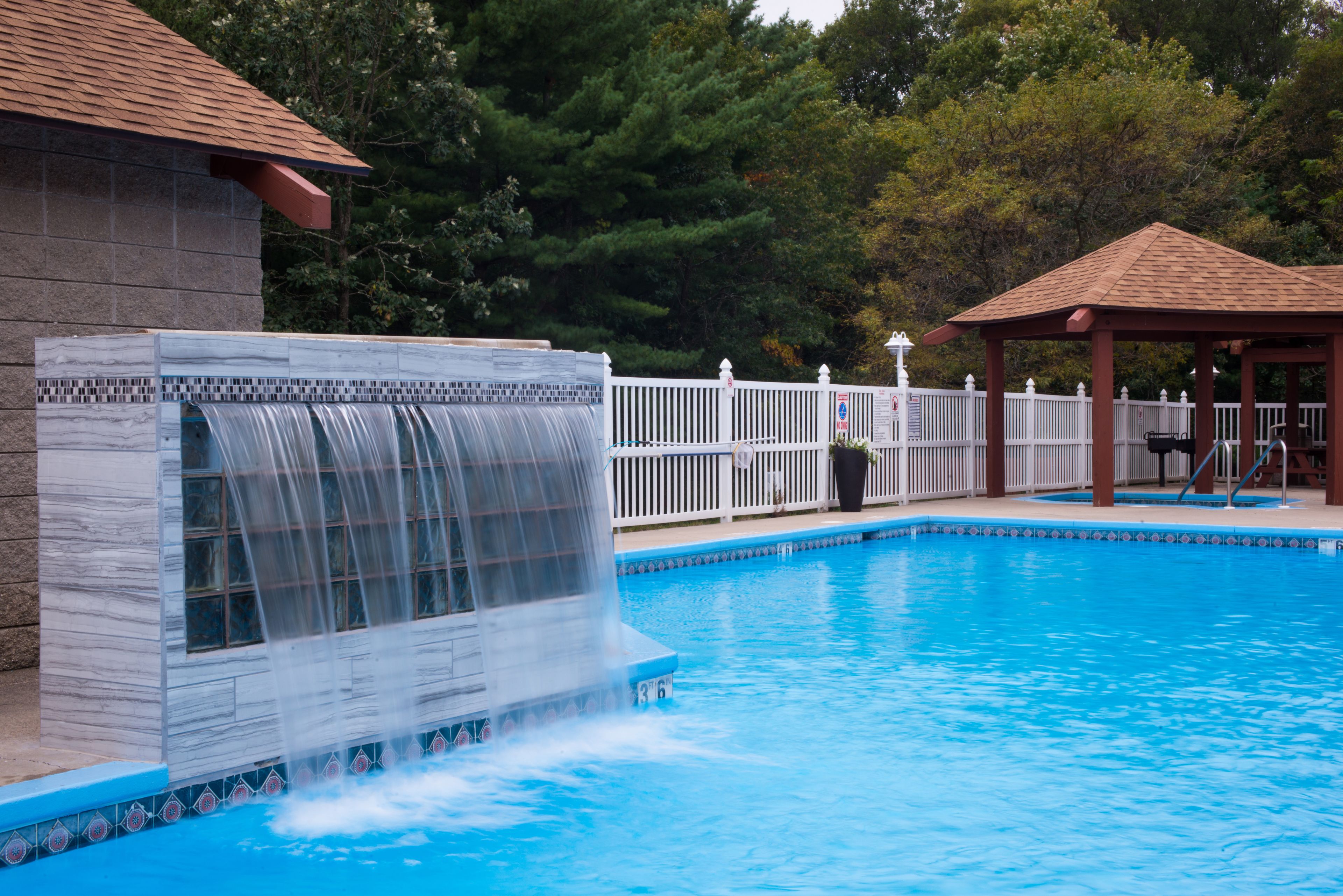 Una piscina con cascata, circondata da un recinto bianco e un gazebo.