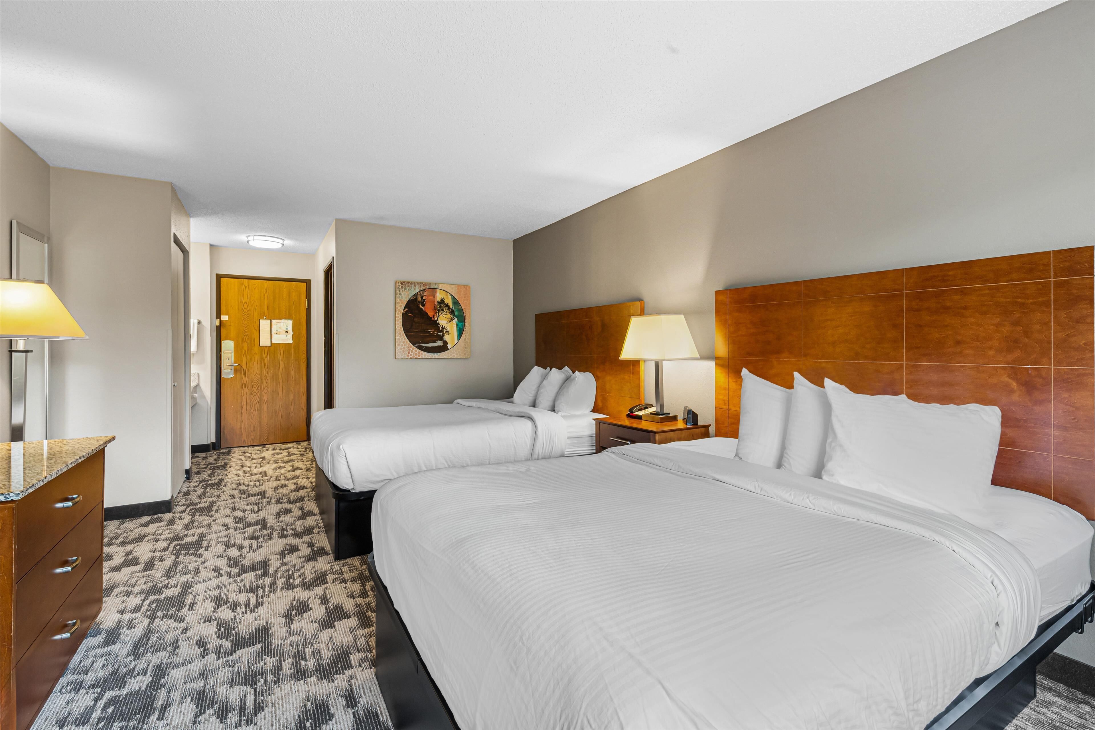 A hotel room with two beds, a wooden headboard, a nightstand with a lamp, a dresser, and a carpeted floor.