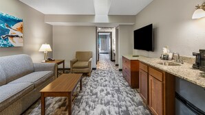 A hotel room with a sofa, armchair, wooden table, kitchenette, and a TV mounted on the wall. - Best Western Ambassador Inn & Suites (Wisconsin Dells)