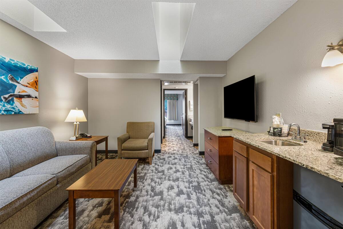 A hotel room with a sofa, armchair, wooden table, kitchenette, and a TV mounted on the wall.