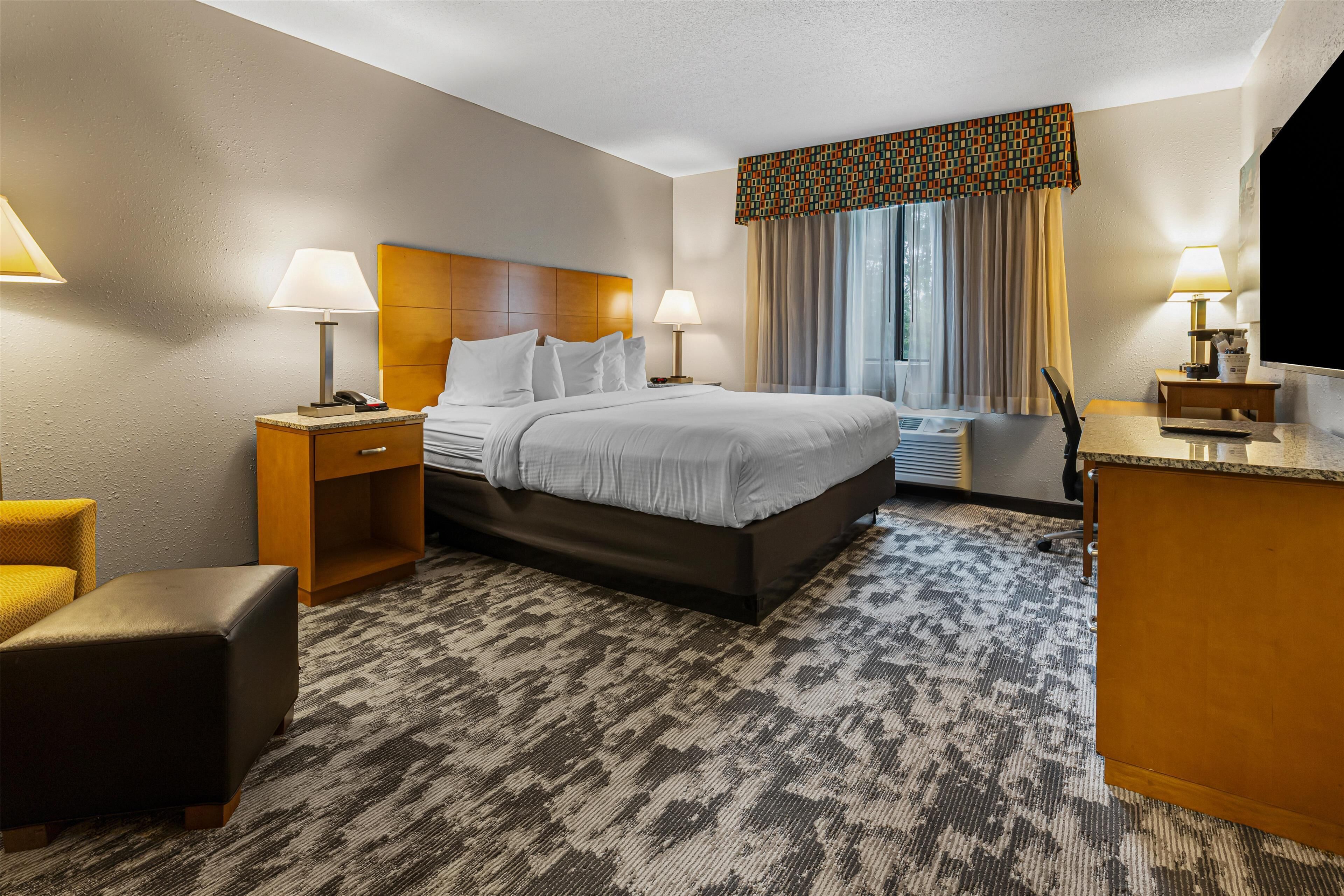 Habitación de hotel con cama, dos mesitas de noche con lámparas, un escritorio con silla, un pequeño reposapiés y un ventanal con cortinas.