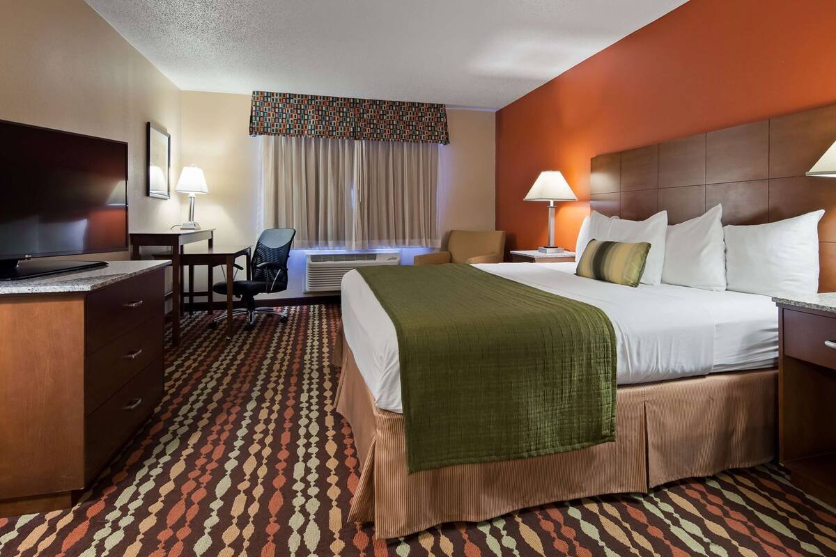 A hotel room with a bed, two bedside lamps, a desk with a chair, a television, and a patterned carpet.