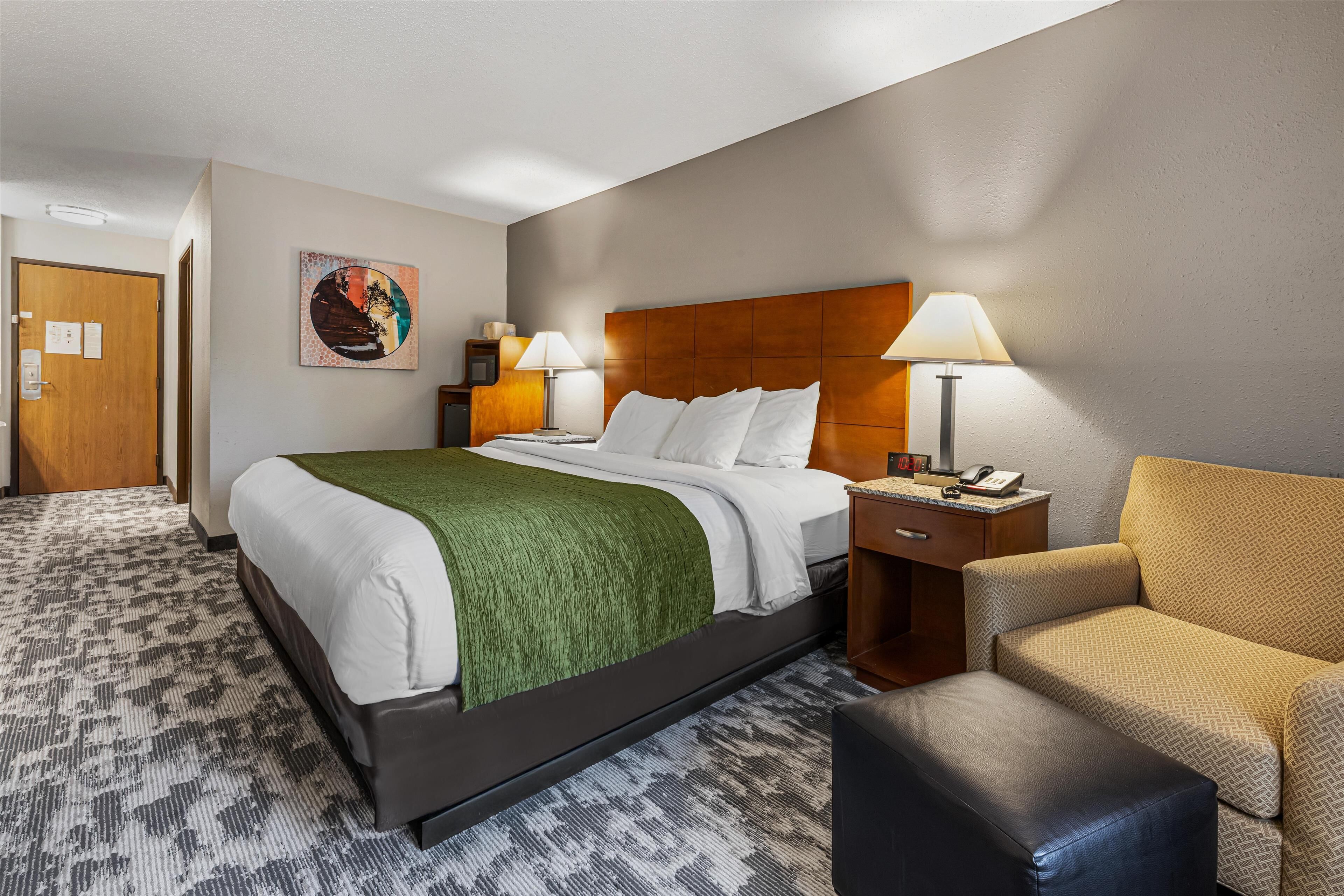 A hotel room with a bed, two bedside tables with lamps, a chair, a desk, and a painting on the wall.