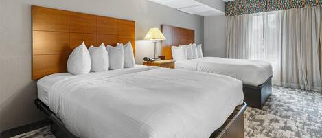 A hotel room with two beds, a nightstand with a lamp, and a window with curtains.