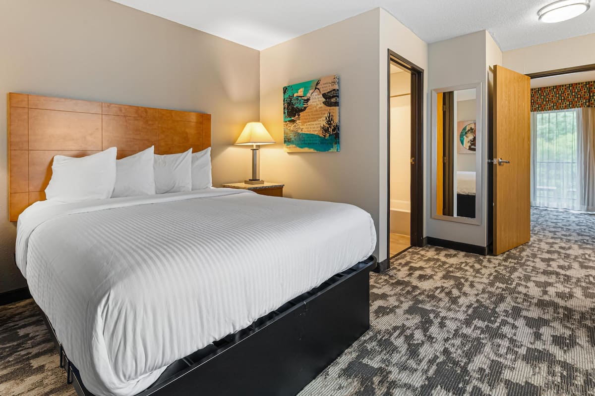 A hotel room with a large bed, a nightstand with a lamp, a painting on the wall, and a door leading to another room.