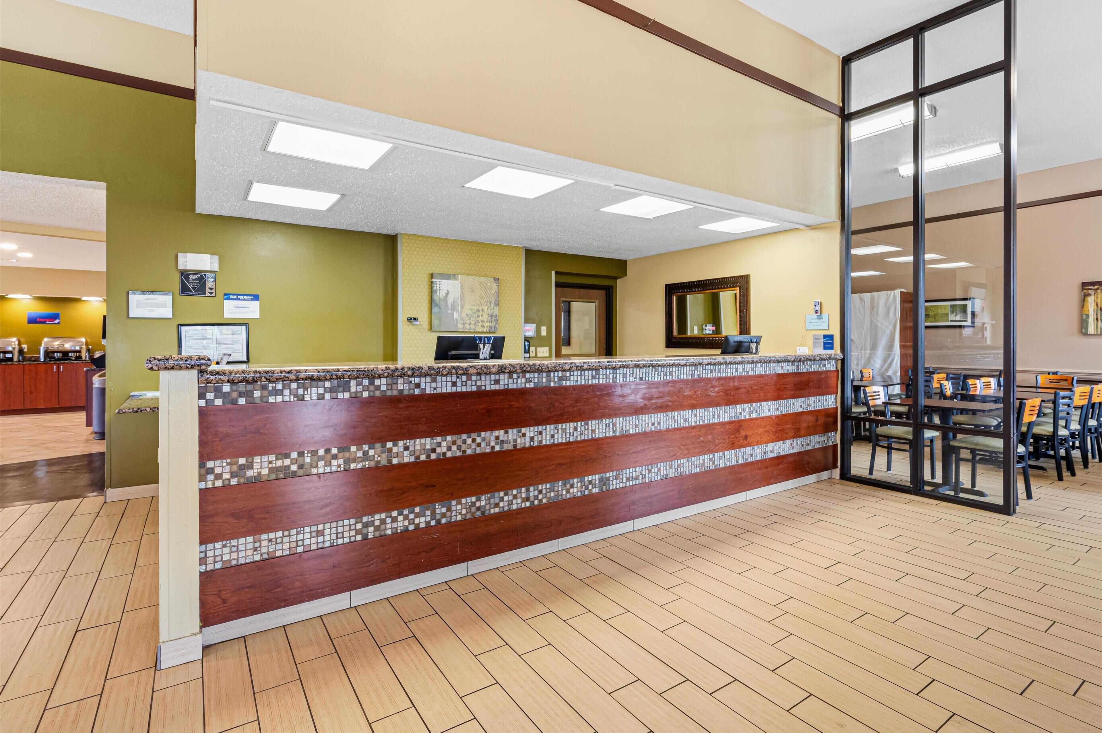 A reception area with a wooden counter, tiled mosaic backsplash, and a glass door leading to a dining area with tables and chairs.