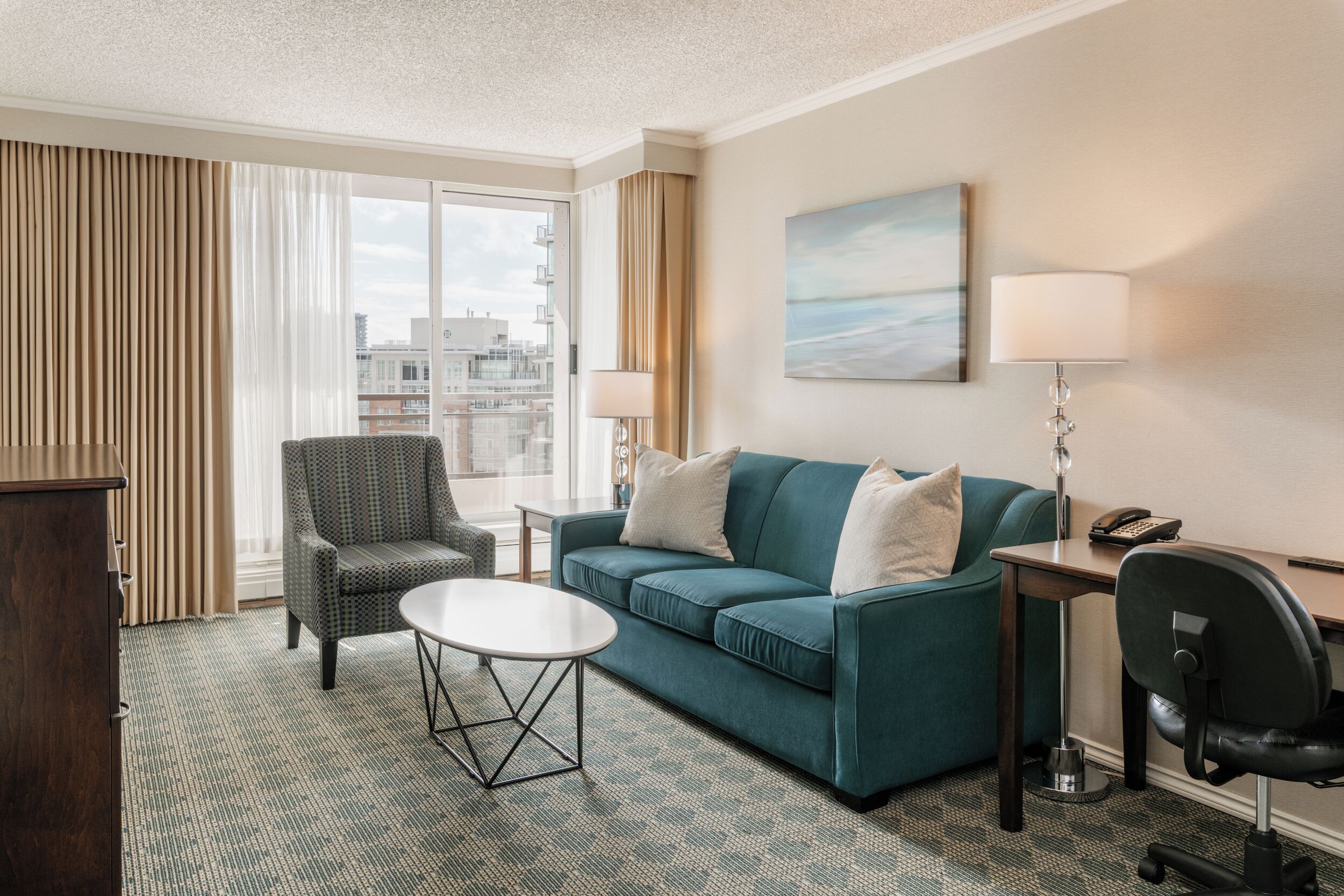 A hotel room with a sofa, armchair, small table, desk, lamp, and a painting on the wall.