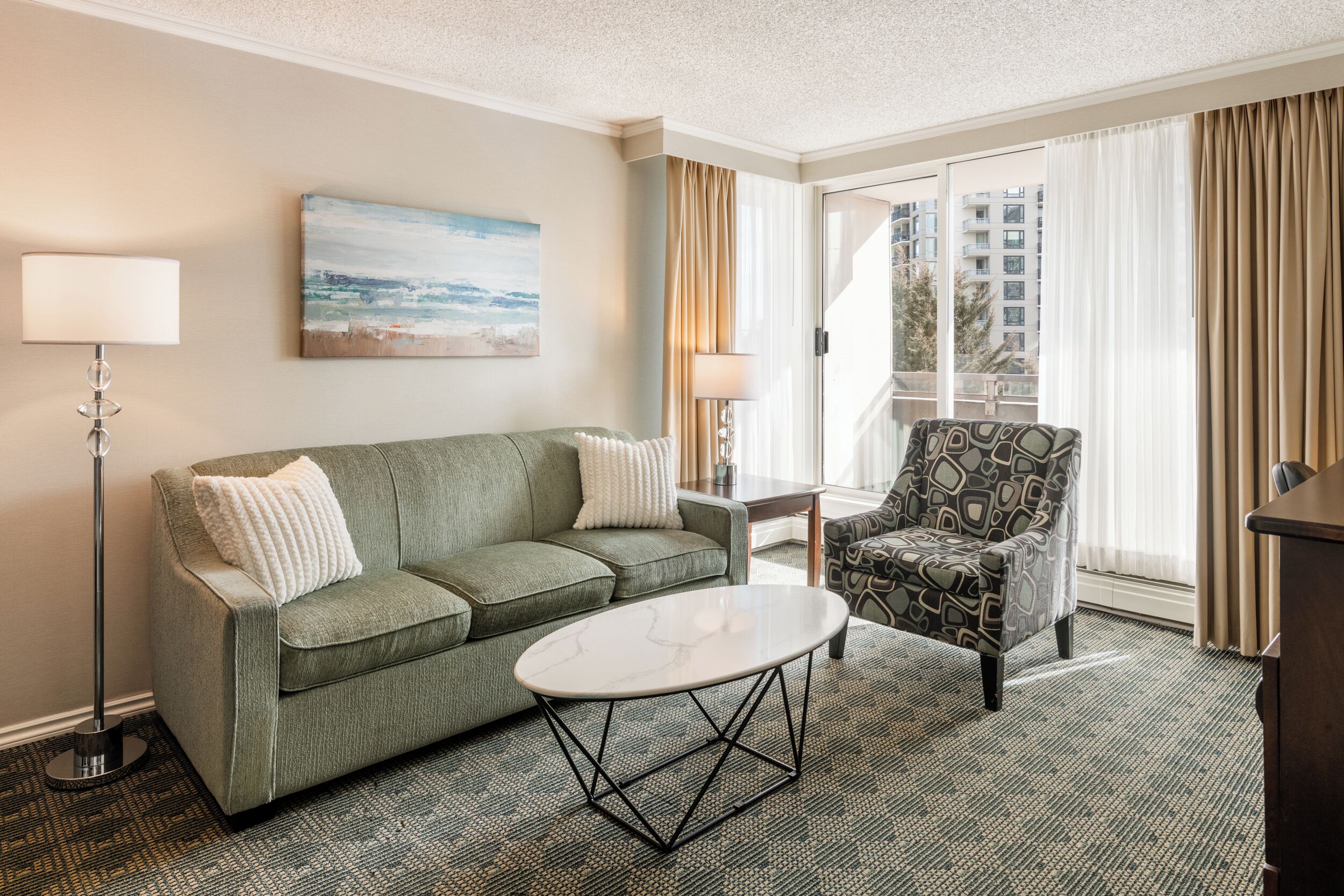 A hotel room with a green sofa, a patterned armchair, a small white table, a floor lamp, and a painting of a beach scene.