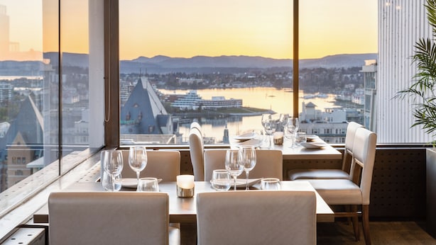 Un comedor con una mesa dispuesta para dos personas, con vista a un paisaje urbano y una extensión de agua durante la puesta de sol.