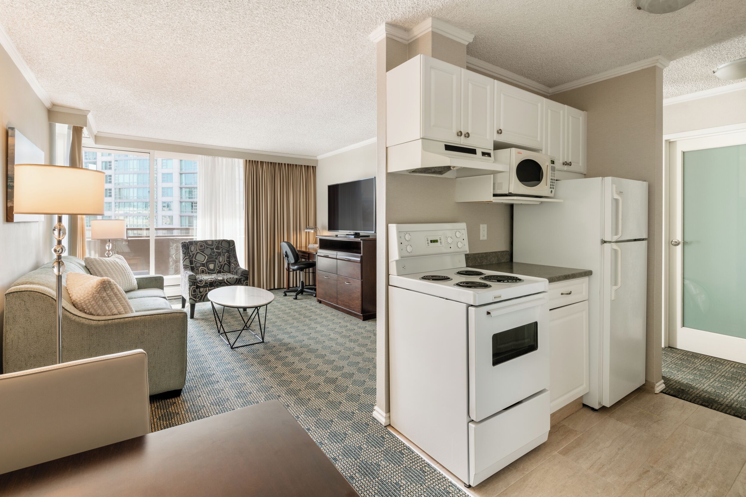 Habitación de hotel compacta con kitchenette, sofá, mesa de comedor y vistas a la ciudad.