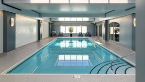 Una piscina interior con agua azul cristalina, techo de cristal y dos sillones de descanso con cojines.