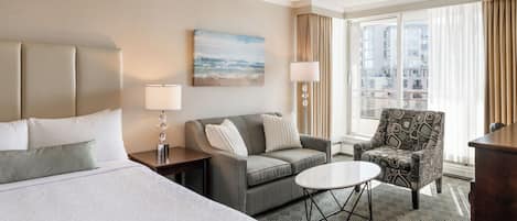 A hotel room with a bed, a sofa, a chair, a small table, and a painting on the wall.
