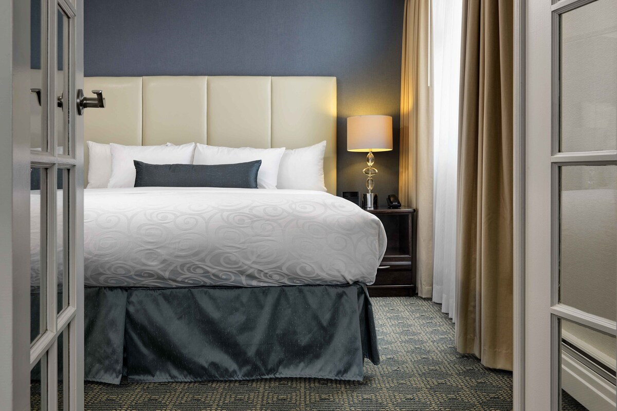 A hotel room with a large bed, a bedside table with a lamp, and a view of the outside through a door.