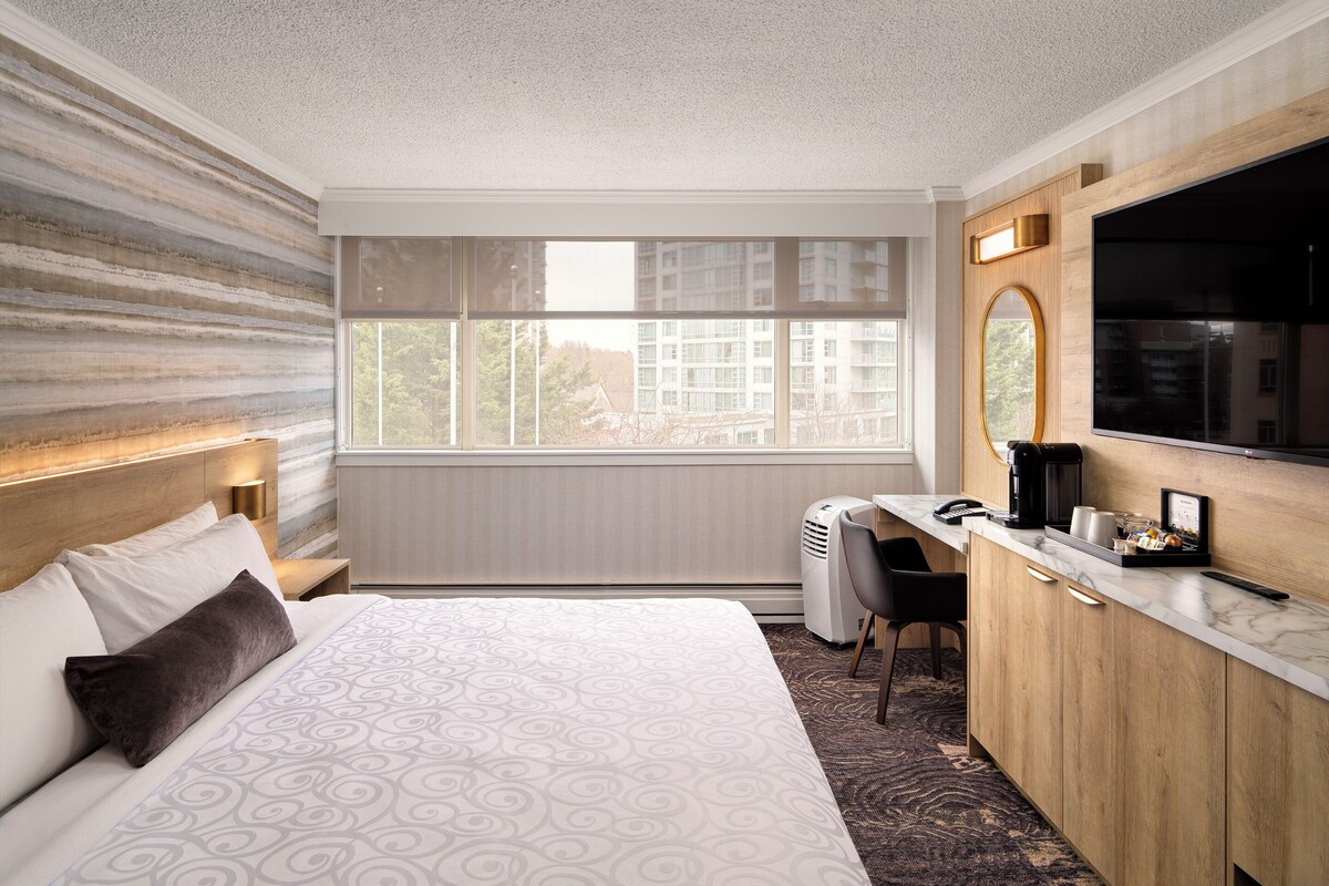 A hotel room with a large bed, a desk, a chair, a TV, and a window with a view of buildings.