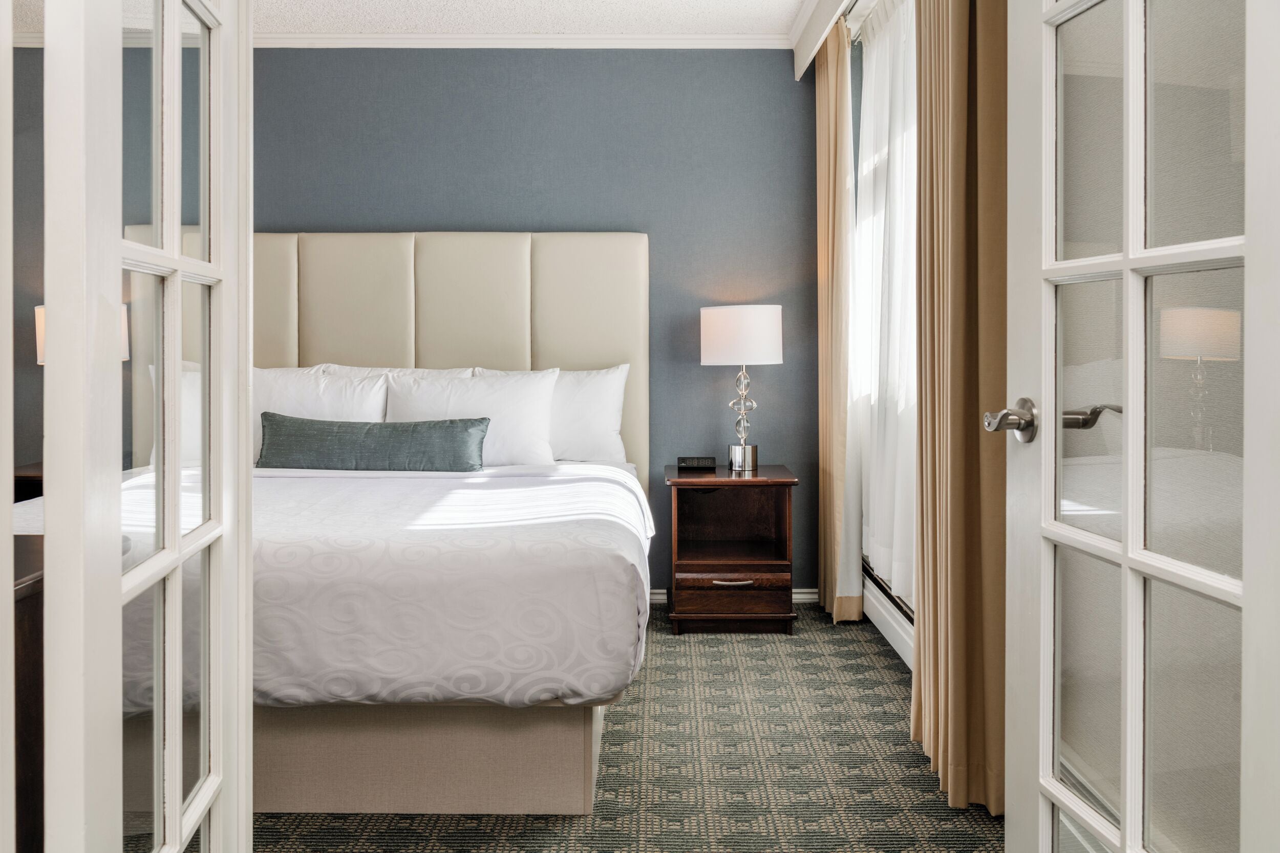 A hotel room with a bed, a nightstand, a lamp, and a view through an open door.