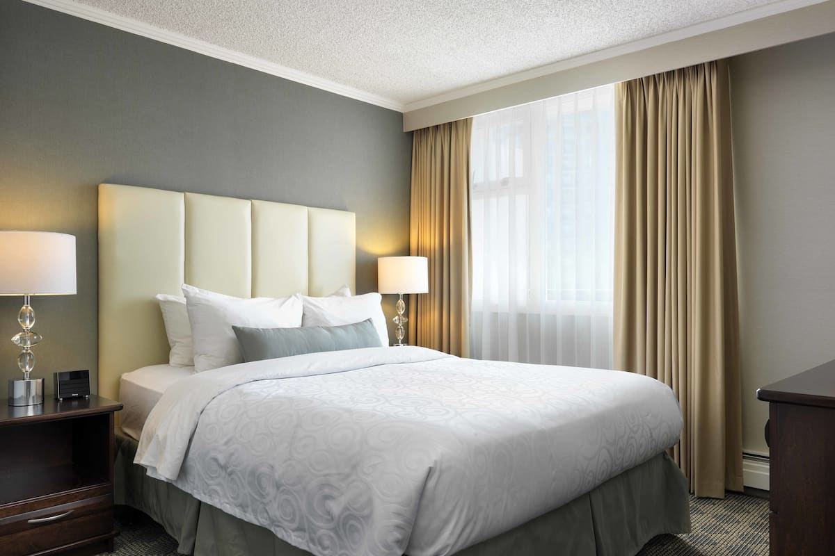 A hotel room with a large bed, two bedside tables with lamps, a headboard, and a view of the outdoors through a window with curtains.