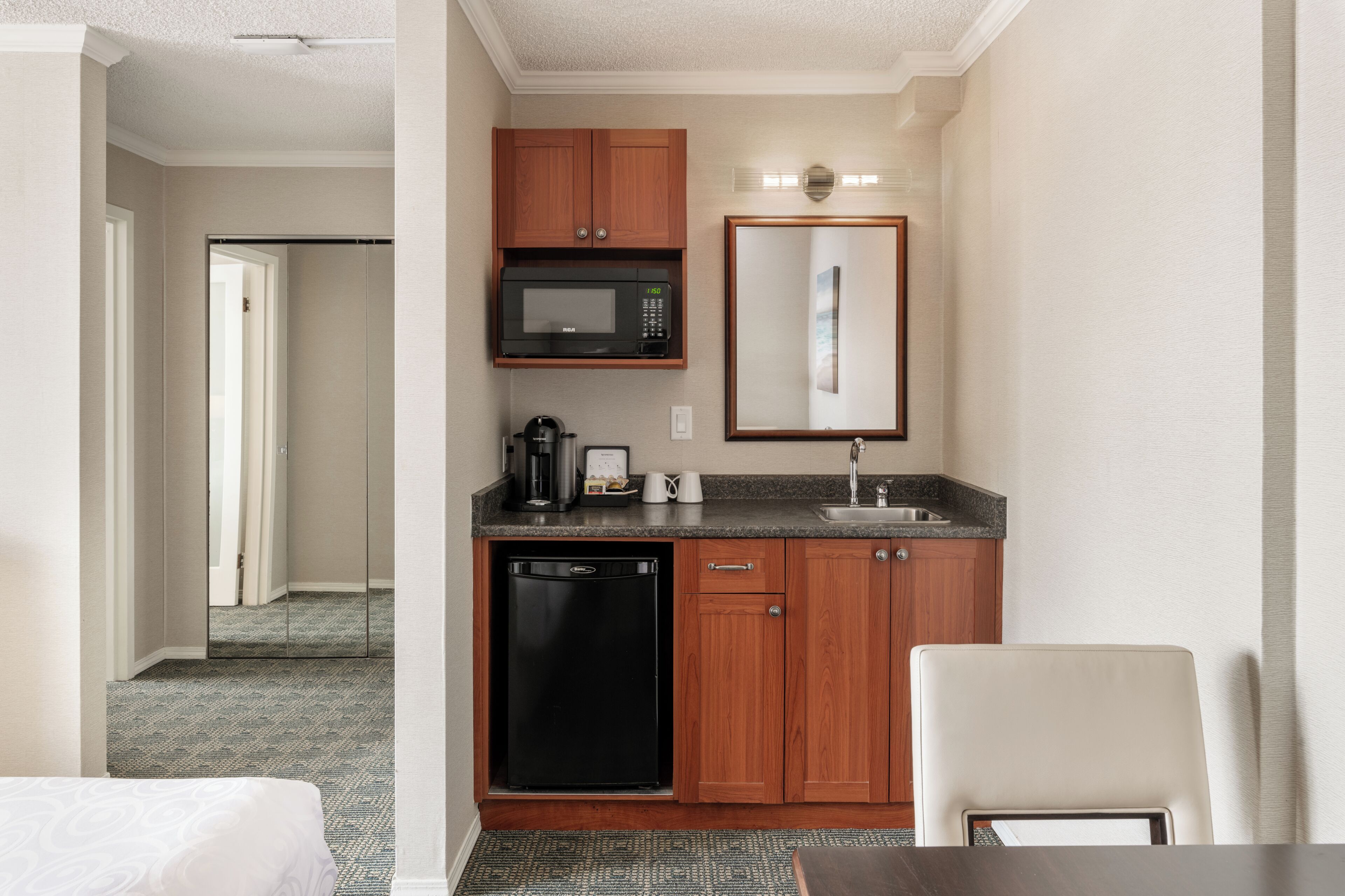 A hotel room with a kitchenette, a microwave, a coffee maker, a dishwasher, a sink, and a mirror.