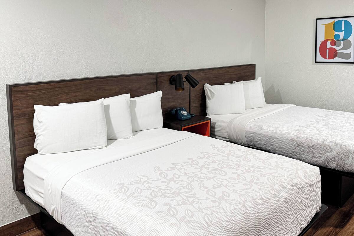 A hotel room with two beds, a nightstand, a lamp, and a framed abstract art piece on the wall.