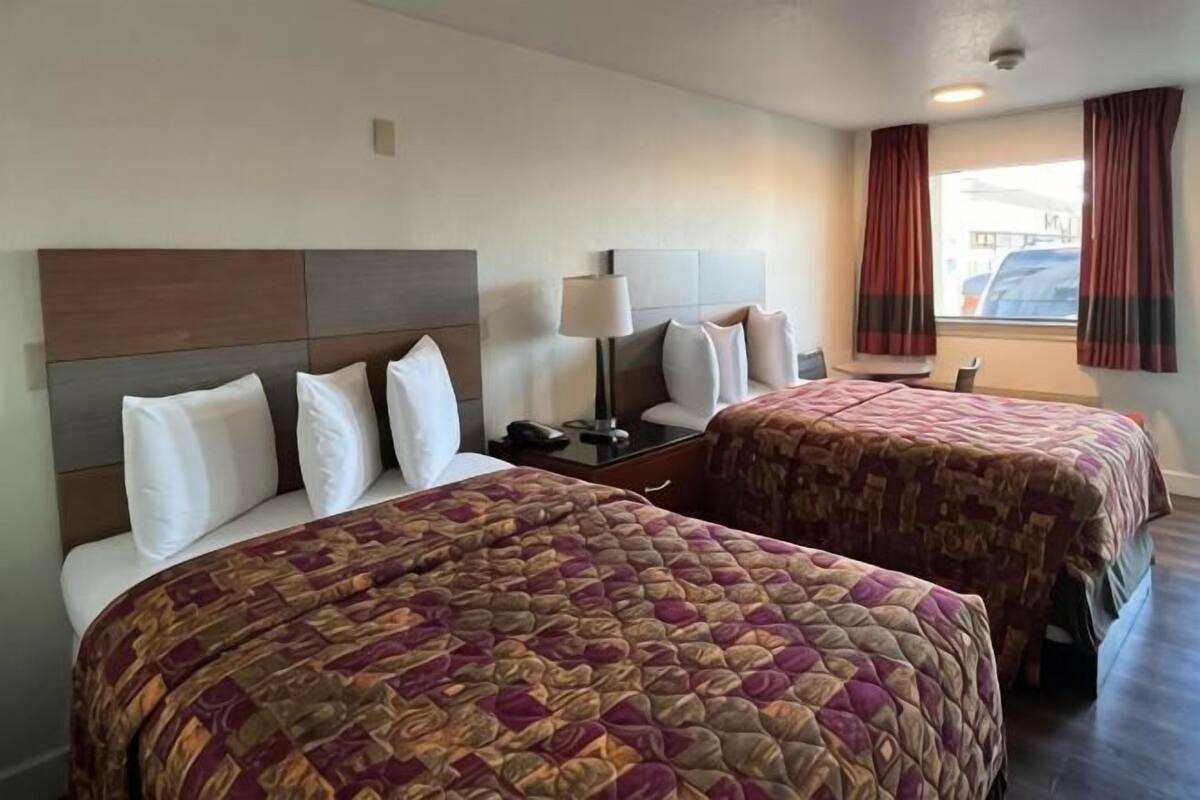 A hotel room with two beds, a nightstand with a lamp, a window with curtains, and a view of the outside.