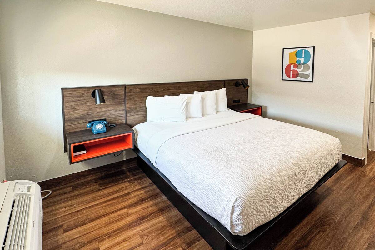A hotel room with a large bed, a bedside table with a lamp, a small shelf, a desk with a chair, and a wall-mounted TV.