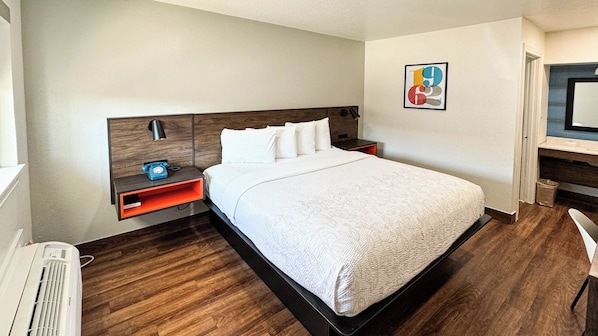 A hotel room with a large bed, a bedside table with a lamp, a small shelf, a desk with a chair, and a wall-mounted TV.