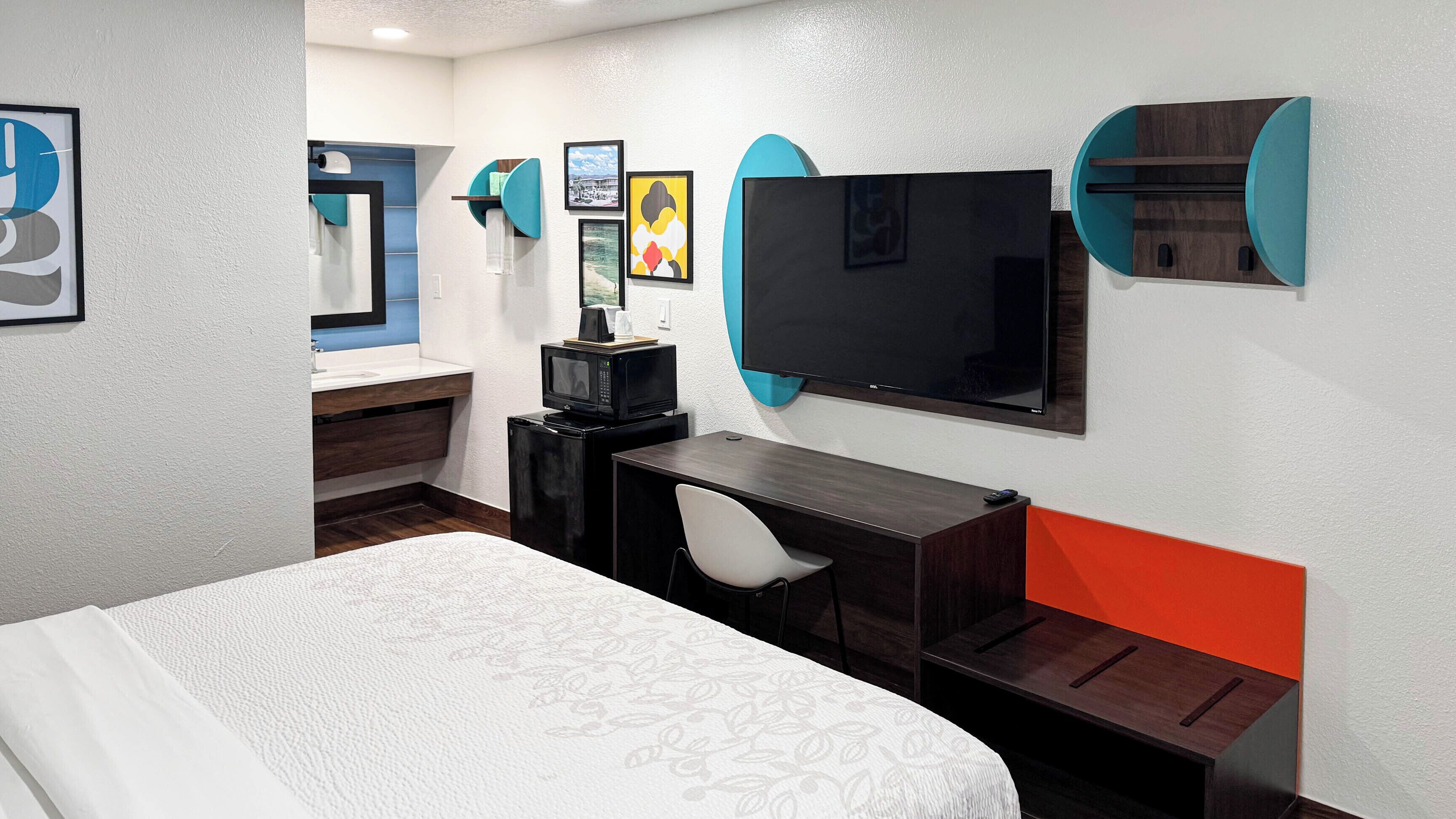 Une chambre d’hôtel comprenant un lit, un bureau avec une chaise, une télévision, un four à micro-ondes et une salle de bain avec un miroir et un lavabo.