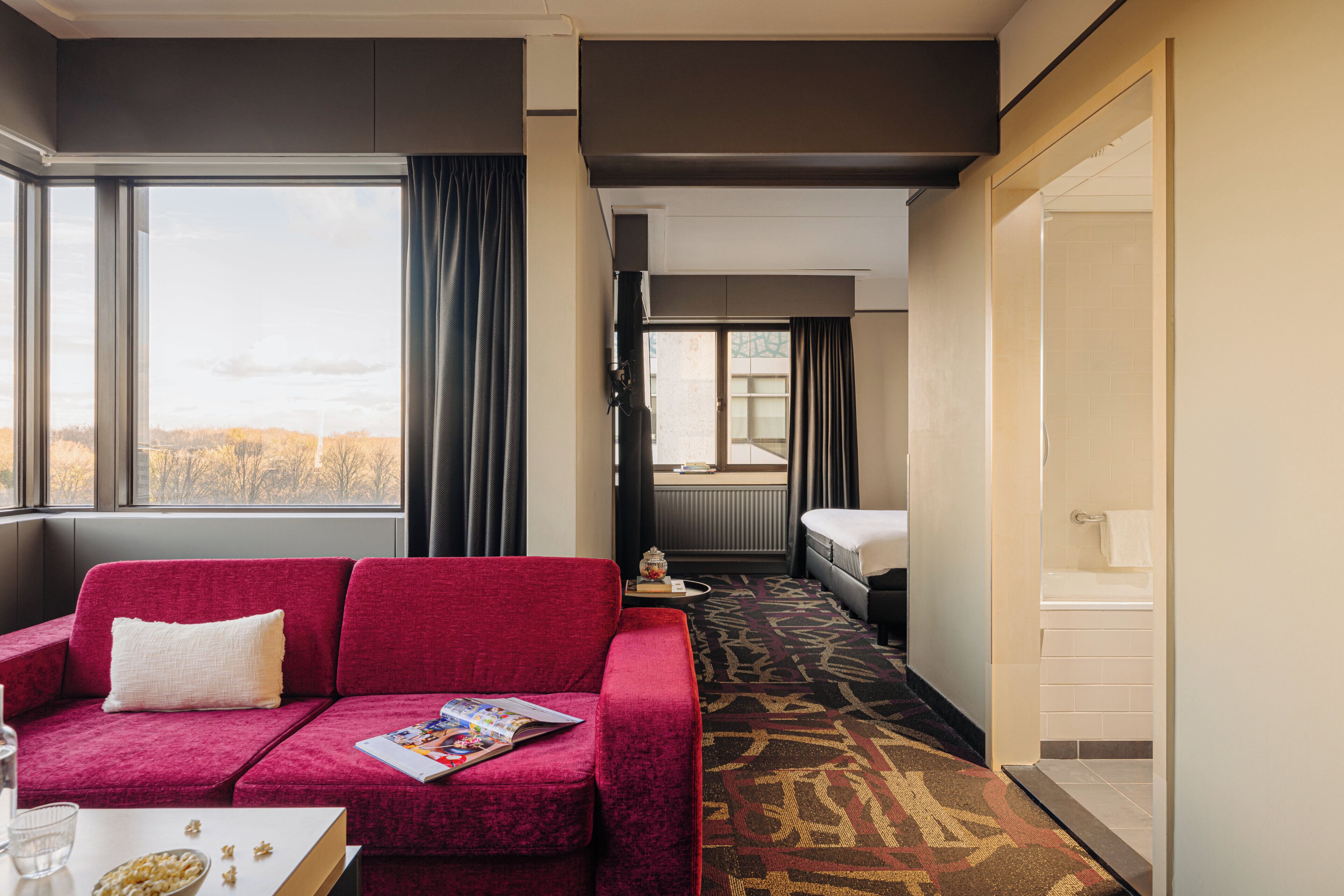 Hotelkamer met een rode bank, een klein tafeltje met een tijdschrift, een raam met gordijnen, een badkamer met badkuip en een kamerbreed tapijt met een patroon.