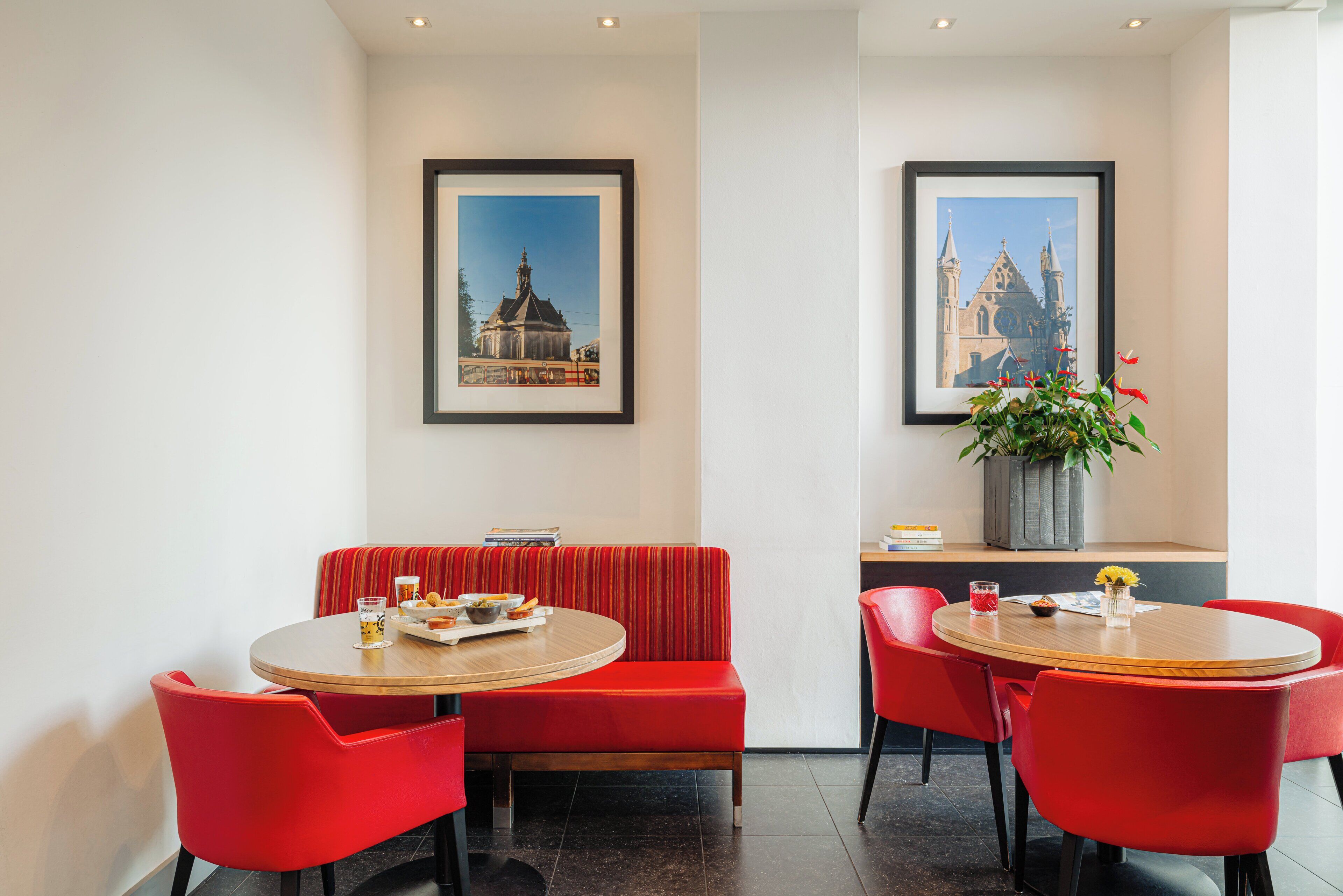 Un coin repas avec une table ronde en bois, un banc recouvert d’un tissu rouge et des chaises assorties. Des tableaux encadrés ornent le mur, ainsi qu’une plante en pot et une petite table sur laquelle est posé un vase de fleurs.