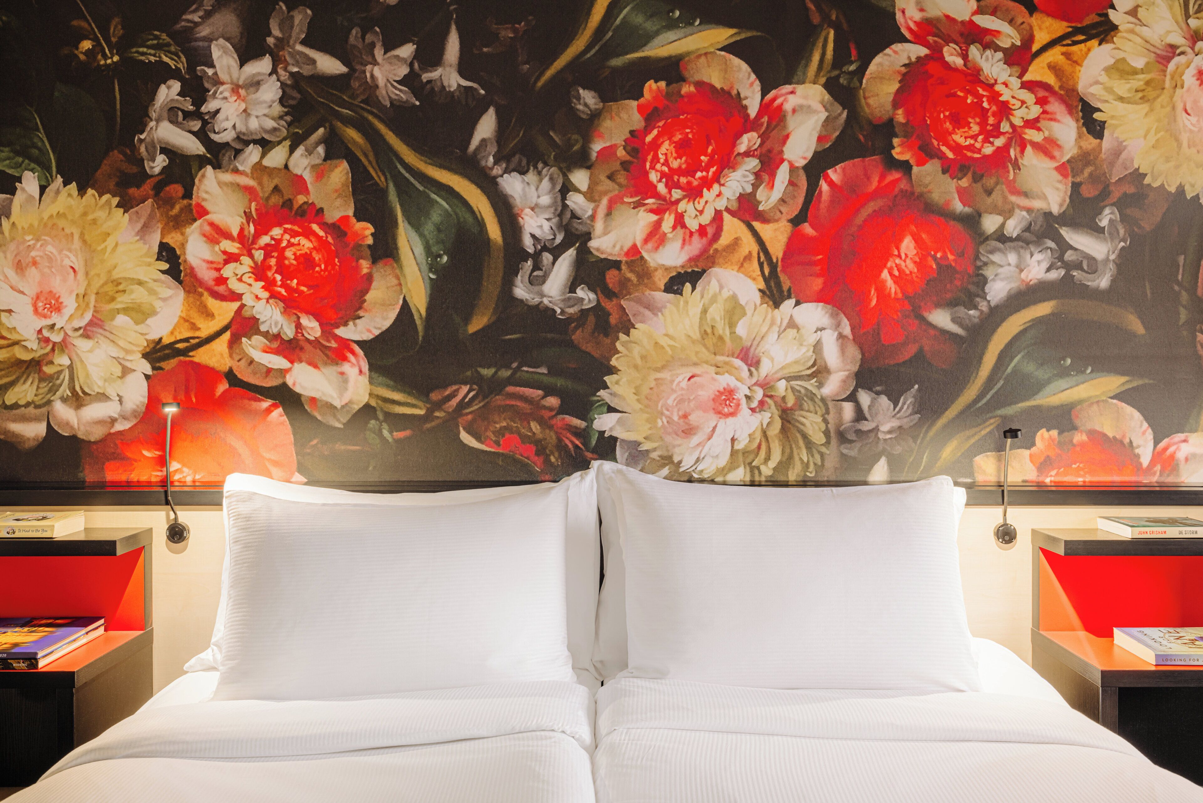 Een hotelkamer met een grote bloemmuur, een bed met witte lakens en rode nachtkastjes met boeken.