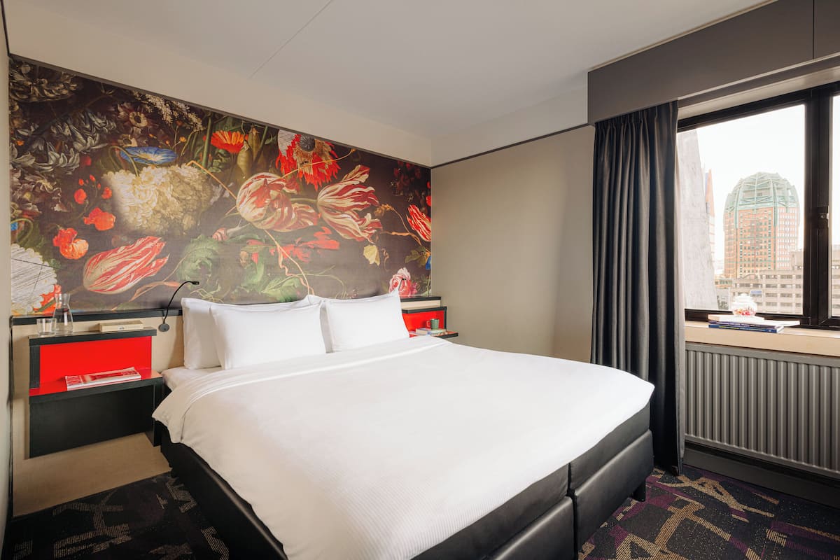 A hotel room with a large bed, a nightstand with a red drawer, a window with a view of a cityscape, and a floral mural on the wall.