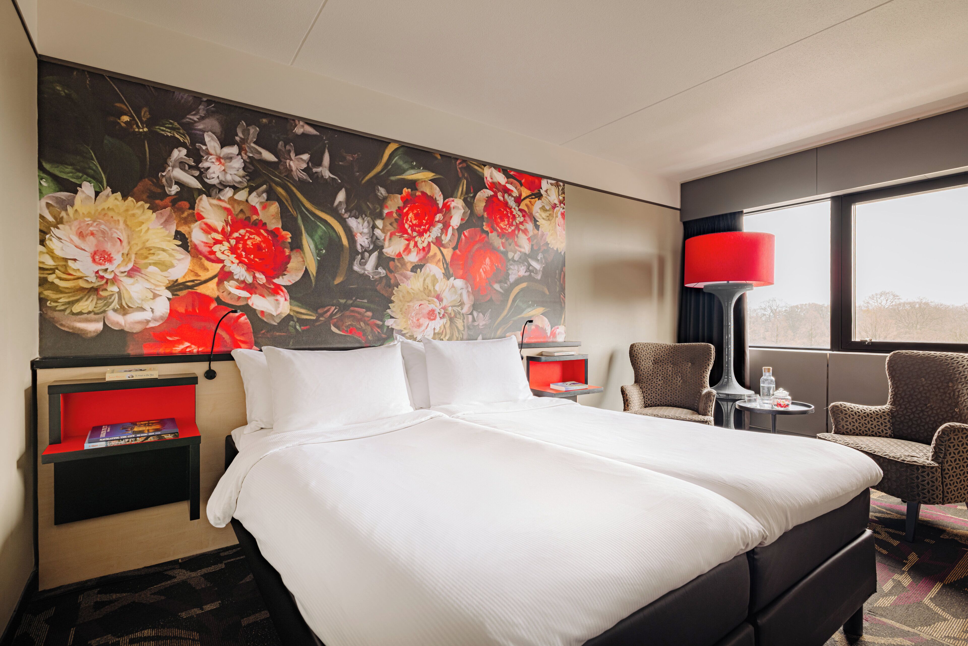 A hotel room with a large bed, a red nightstand, a red lamp, a chair, and a large floral painting on the wall.