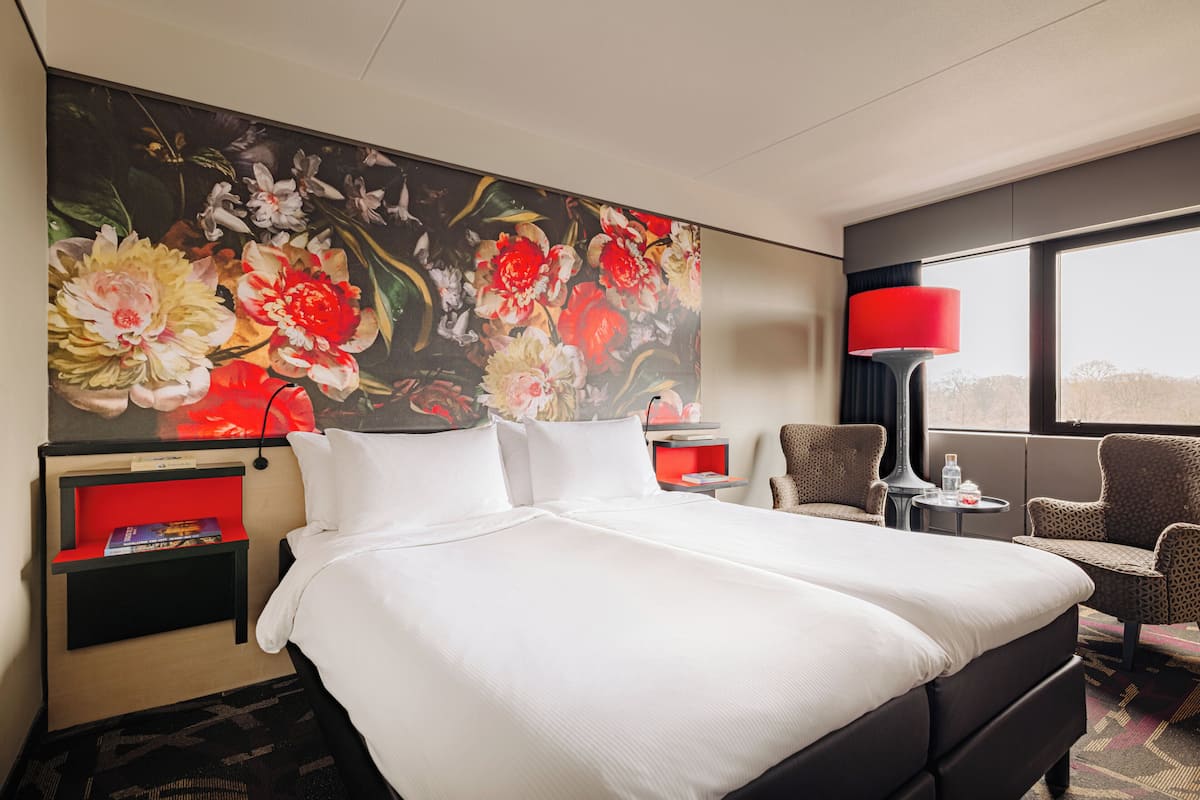 A hotel room with a large bed, a red nightstand, a red lamp, a chair, and a large floral painting on the wall.