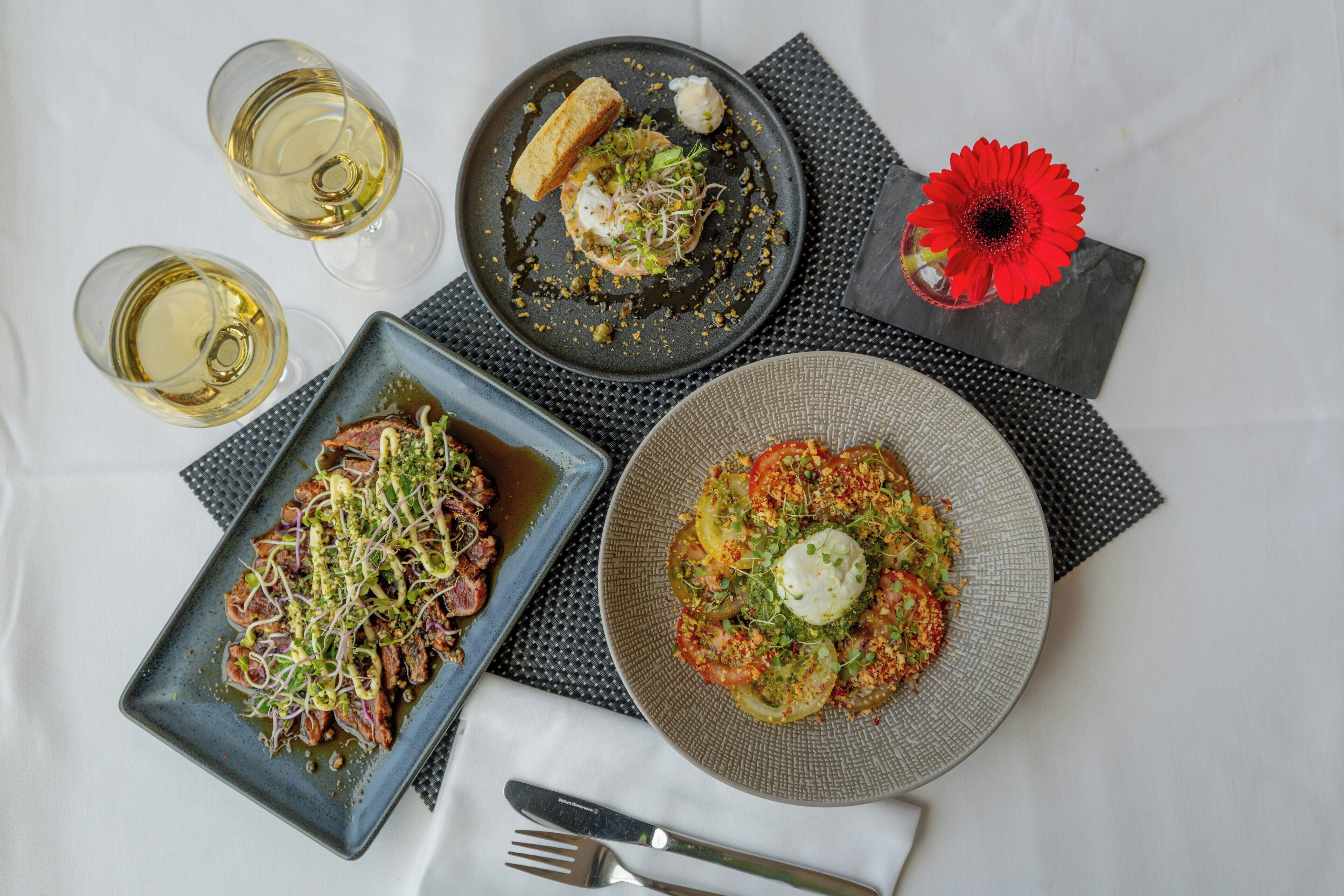 A table with three plates of food, two glasses of white wine, and a red flower on a napkin.
