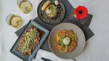 A table with three plates of food, two glasses of white wine, and a red flower on a napkin.