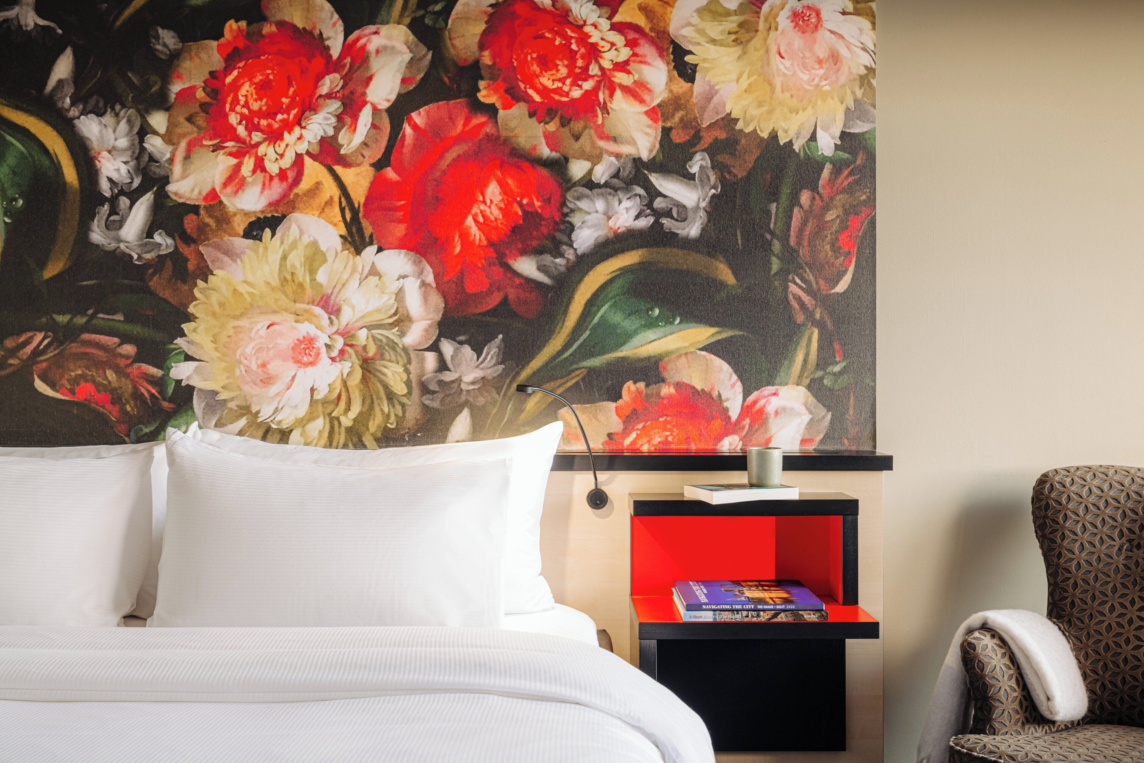 Hotelkamer met een bed, nachtkastje met lamp, stoel en wand met bloemschilderij.