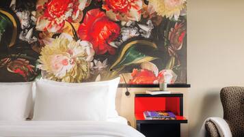 Una habitación de hotel con una cama, una mesita de noche con lámpara, una silla y un cuadro floral en la pared.