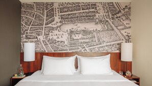 A hotel room with a large map mural on the wall, a wooden headboard, two white pillows, a bedside table with a lamp, and a glass of water. - Leonardo Hotel Den Haag Babylon (The Hague)