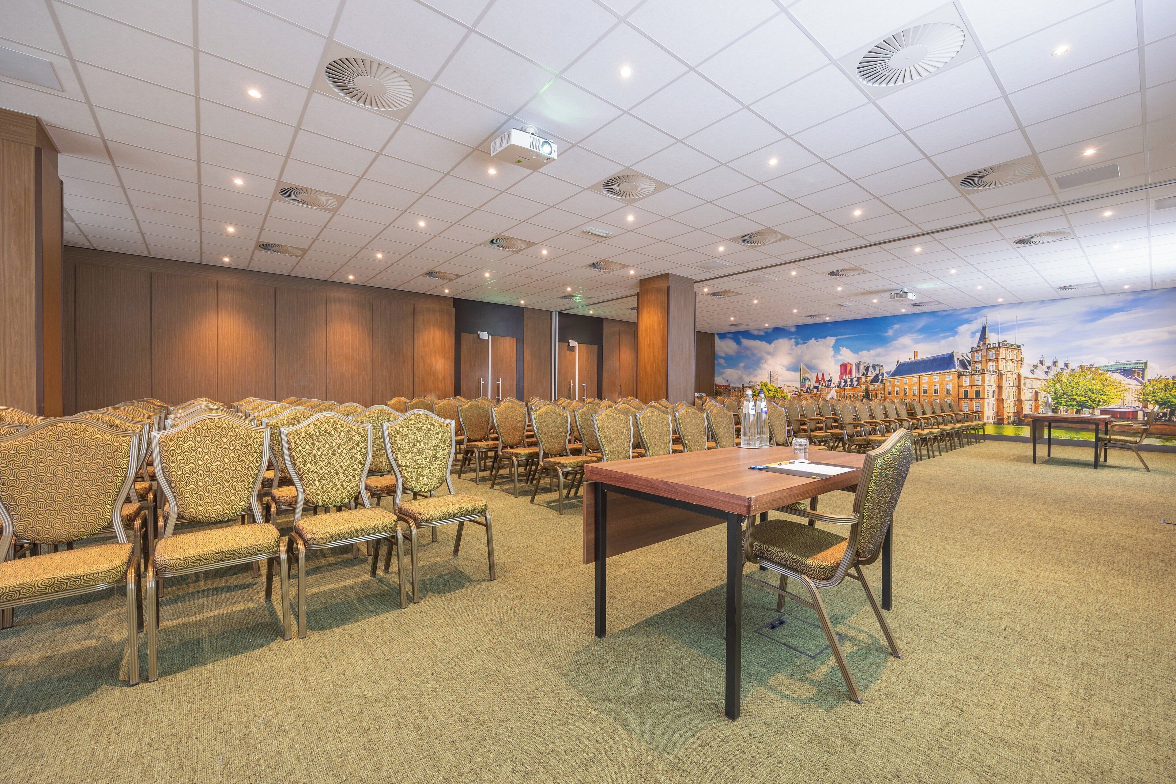 Une salle de conférence avec des rangées de chaises, une longue table et une fresque représentant un paysage urbain sur le mur.