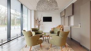 Un área de espera moderna con sillones verdes, una lámpara decorativa en el techo y un amplio ventanal con vistas al exterior.