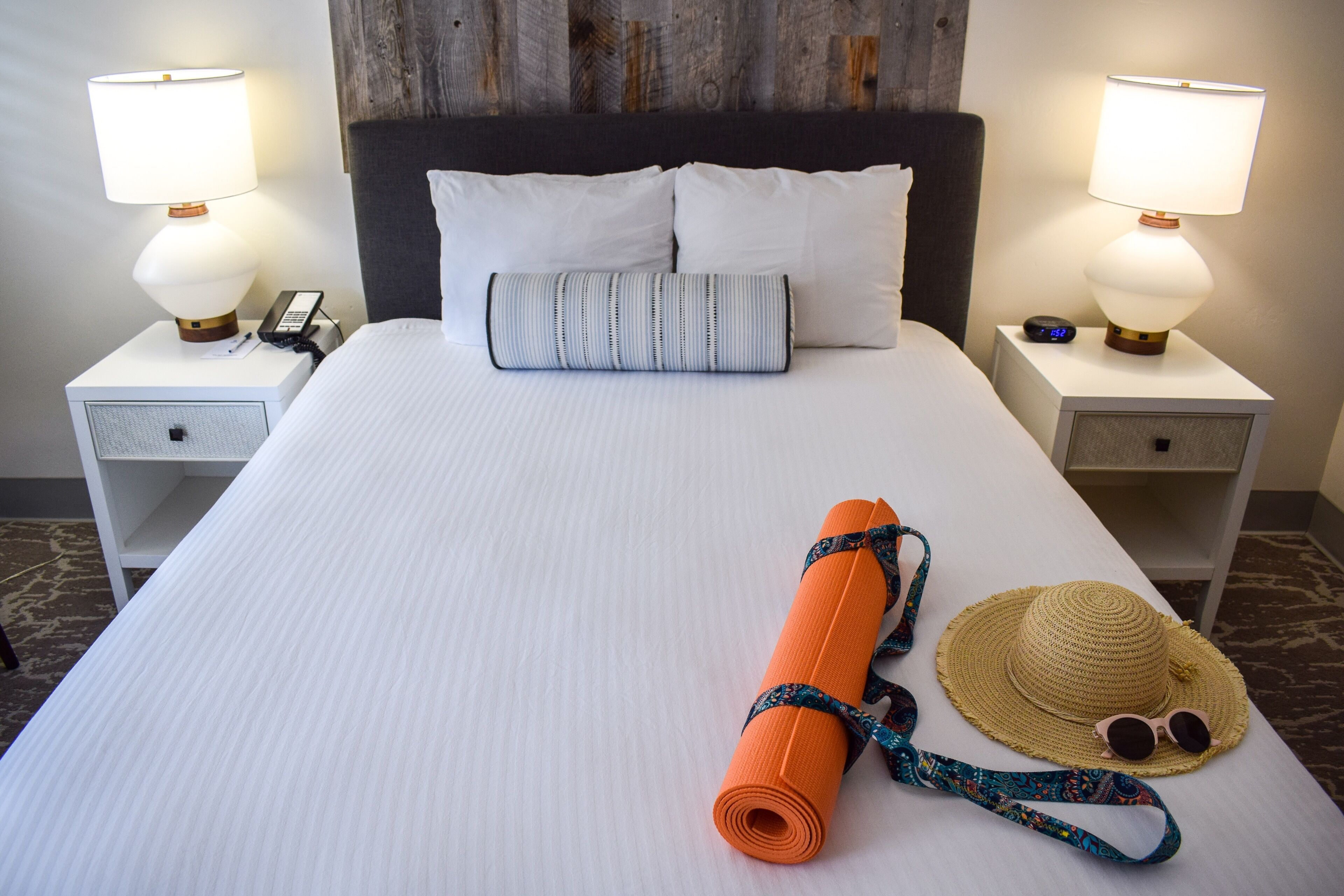 A neatly made bed with white linens, two bedside lamps, a small bedside table with a phone, and a rolled-up orange yoga mat with a matching strap.