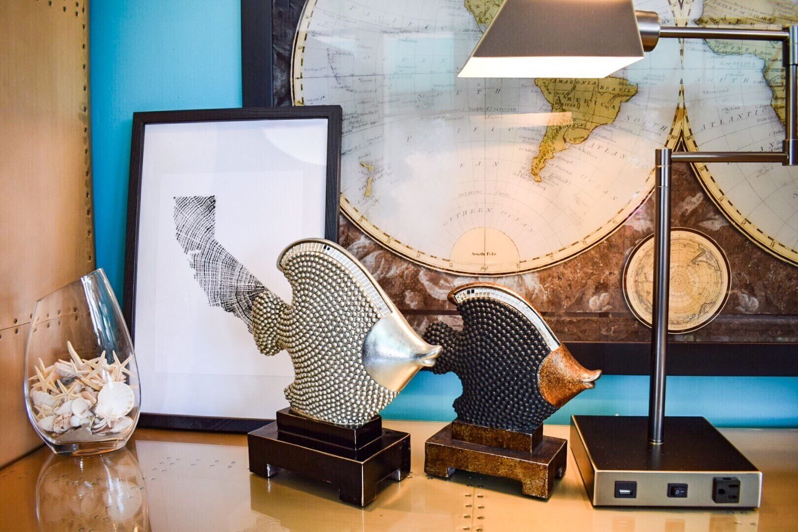 A desk with a framed map, a decorative sculpture, a glass container with seashells, and a lamp.