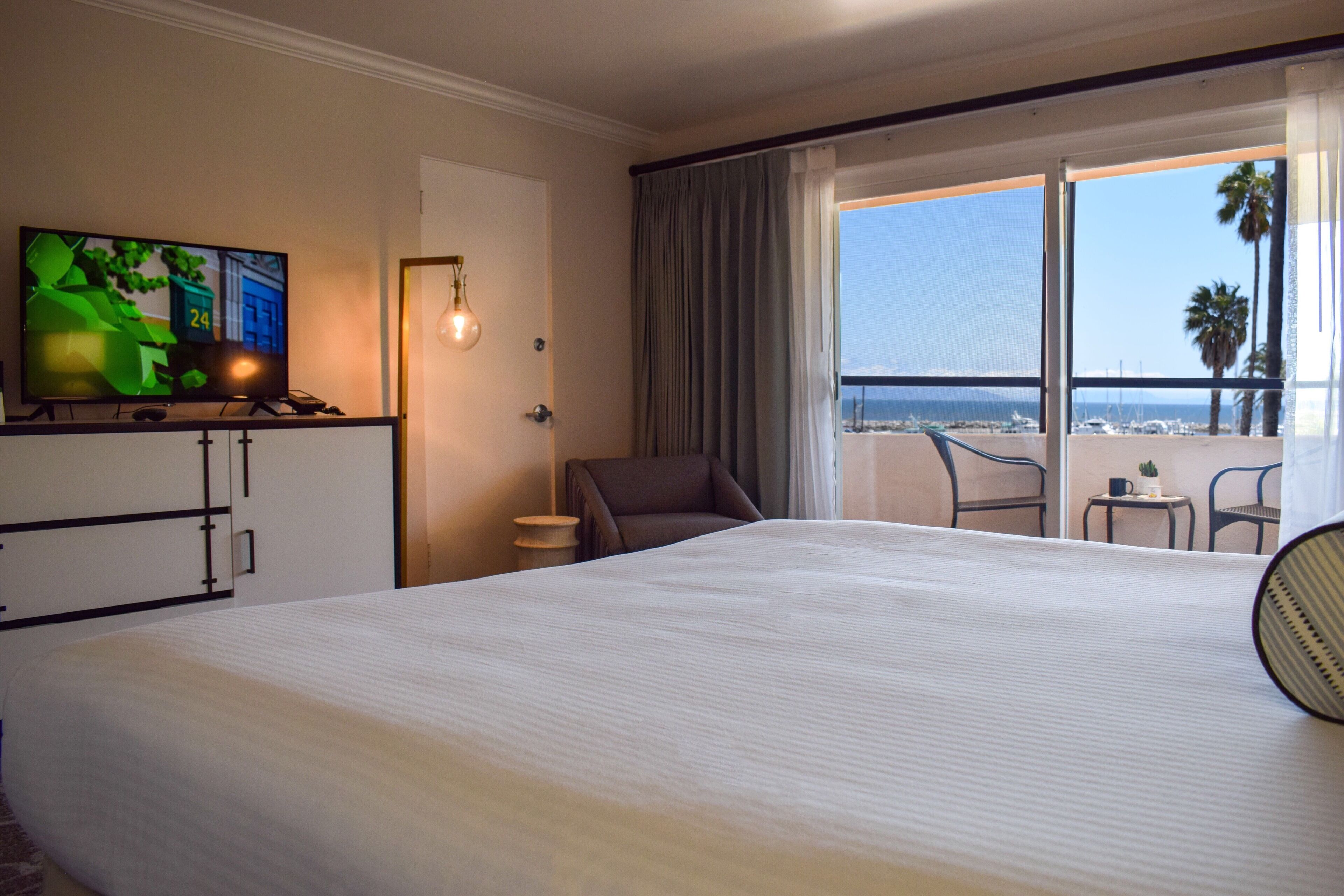 Habitación de hotel con una cama grande, un televisor sobre un soporte, una silla, una mesita y vistas a la playa a través de un balcón.
